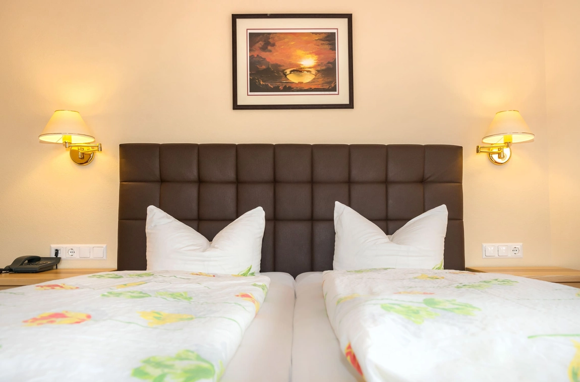 Doppelbett mit floralem Bettbezug, zwei Wandlampen und Sonnenuntergangsbild an der Wand über dem gepolsterten Kopfteil.
